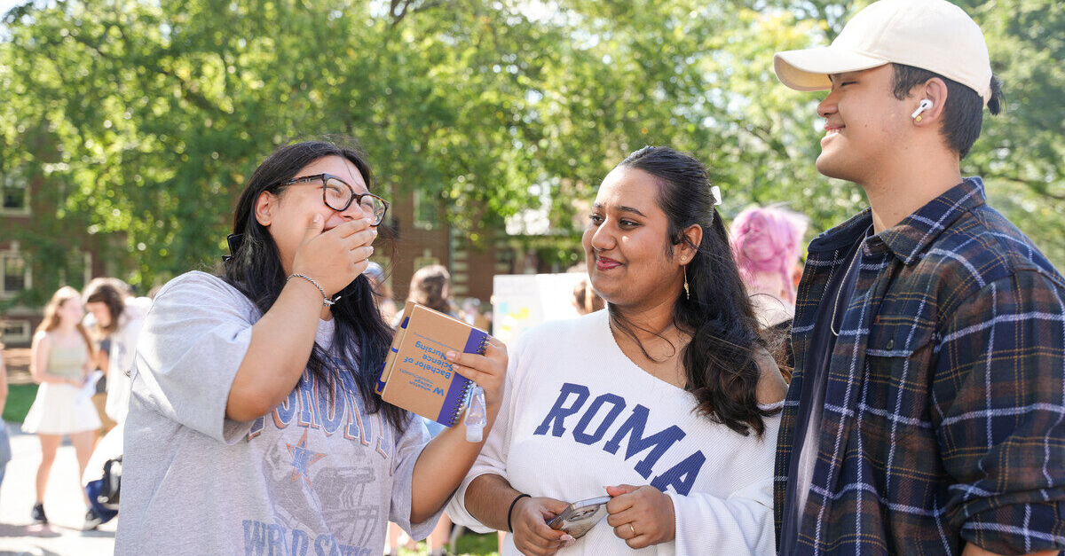 Three students laughing and smiling in an outdoor setting on a bright, sunny day