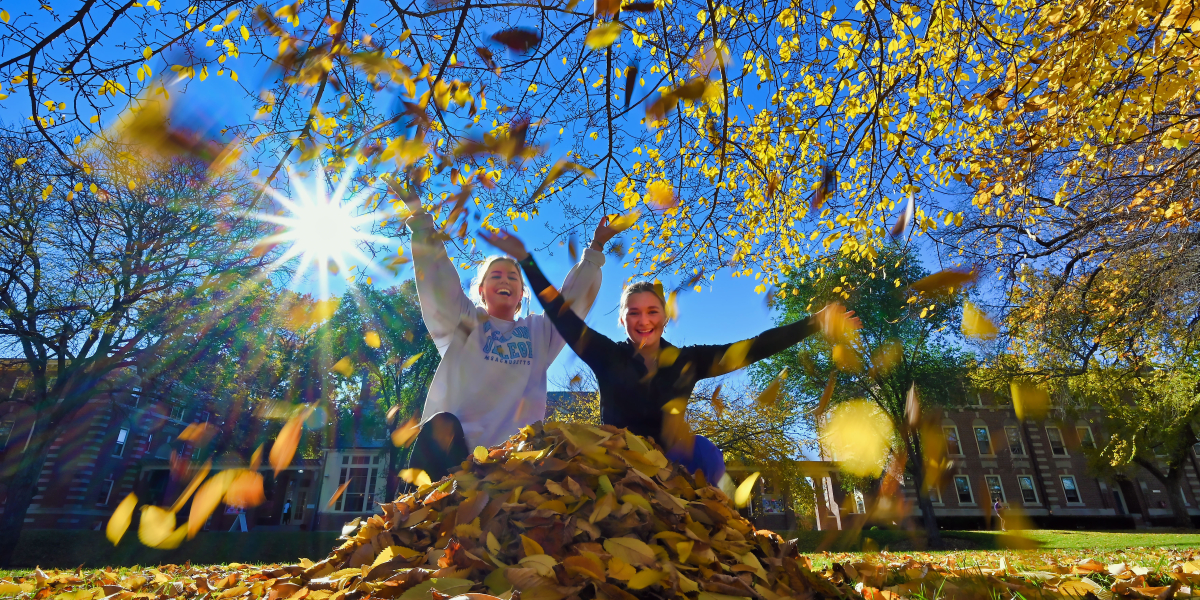 Students toss colorful leaves in the air on a fall day on campus at Wheaton College